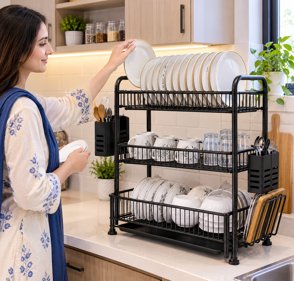 A woman in a kitchen places white plates onto a black three-tier wire dish rack filled with dishes, mugs, and utensils. The scene is set in a bright, modern kitchen with light wood cabinets and white tile backsplash.
