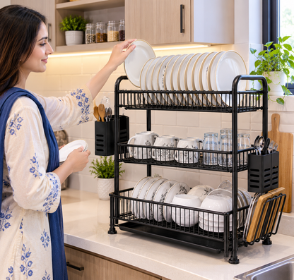 A woman in a kitchen places white plates onto a black three-tier wire dish rack filled with dishes, mugs, and utensils. The scene is set in a bright, modern kitchen with light wood cabinets and white tile backsplash.